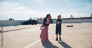 Young attractive woman with baby boy exploding firecracker confetti in slow motion on rooftop on sunny day. Carefree family having fun together, cheerful mom and son on the roof parking