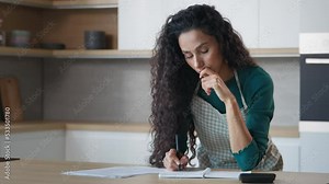 Concentrated hispanic woman wife young mom stand in modern kitchen make notes at copybook write recipe grocery list calculate domestic utilities use calculator count bills puzzled by monthly expenses