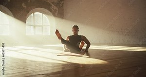 A man in a black sports summer uniform is doing a scissors exercise in a sunny brick hall. Fitness classes on a special mat on the floor in the gym