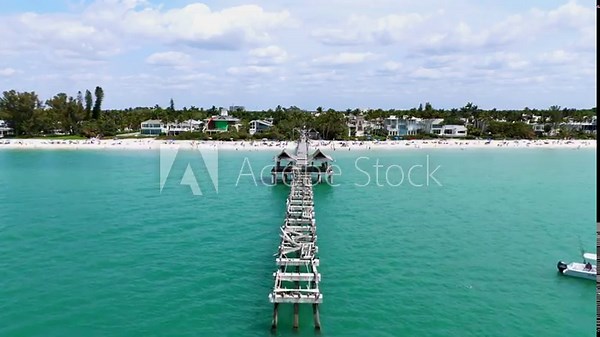 Cement pier with white railing extending from beach into water. Crystal-clear turquoise ocean reflecting sky. White sandy beach bordering lush green landscape and buildings