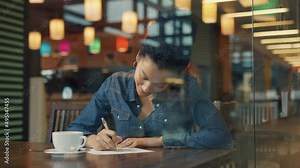 View through a glass window of a young African American woman dreamily thinking about something and then squeaking on a piece of paper. Woman enjoying coffee in a mall cafe. Close. Slow motion.
