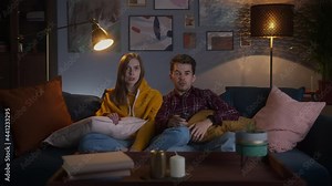 Young couple watching film or entertainment show on TV, next gen 2020, friends switching channels with the remote control, sitting on sofa at home, having fun during coronavirus lockdown.