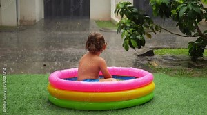 Toddler sits in small inflatable pool under pouring tropical rain, facing away. Contemplating rain in stillness, everything around is soaked, and warm downpour brings refreshment on hot day. Stock Video