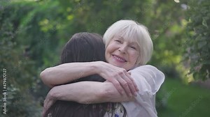 Happy senior elegant woman hugging young granddaughter in slow motion smiling looking at camera. Portrait of positive beautiful Caucasian retiree meeting relative in spring summer park posing