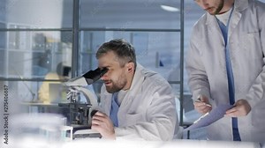 Medium shot of Caucasian male junior researcher, wearing lab coat, coming up to middle-aged project leader, who is looking into microscope, and demonstrating him report sheet with experiment results Stock Video