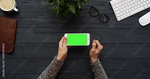 Top view on the black office desk and white smartphone with green screen and female hands taping on it. Horizontal. Office stuff beside. Chroma key.