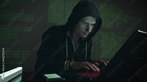 A male hacker in a hood typing program code while committing a cybercrime hacking a system firewall on the background of screens in the dark under neon light. Cyber security concept