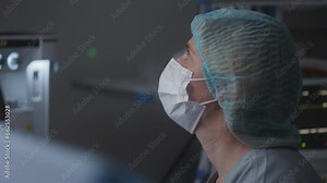 Female adult medic in medical hat and mask sits in modern operating room and talks with surgeons. Nurse looks at electrocardiography monitor and checks condition of patient during difficult surgery.