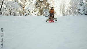 Two male friends having fun in forest on winter day: young cheerful man lying on stomach on sled and riding down the hill while his friend holding his legs and running behind