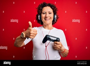 Middle age curly hair gamer woman playing video game using joystick and headphones happy with big smile doing ok sign, thumb up with fingers, excellen Stock Photo - Alamy