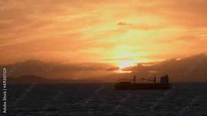 A small cargo ship is silhouetted against a setting sun at sea. The yellow clouds contrast against the ocean foreground.