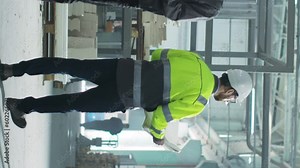 Vertical shoot. Full height engineer is holding blueprint. Builder dressed up safety clothes and hard hat. Male at backgrounf of equipment and work process. Worker inspects construction site.
