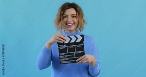 Happy young woman movie clapper slow motion in hands in blue sweater on blue background. Assistant director holds clapper board and claps his hands, smiling, looking at camera. Film industry. Monotone