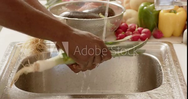 African American man holding leek at sink, rinsing, peeling leaves under running tap to remove dirt