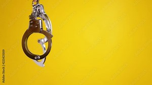 Shaking a pair of shiny metal handcuffs over a yellow background, room for copy-space on the right. Symbolic shot: crime, punishment, menace, fetish, sexuality, pleasure.