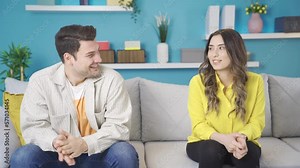 First date. Shy, young attractive lovers looking at each other, excited and happy. Happy and excited couple sitting on sofa at home, first date excitement and shyness.