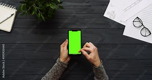 Top view on the black office desk and black smartphone with green screen and female hands taping on it. Vertical. Office stuff beside. Chroma key.