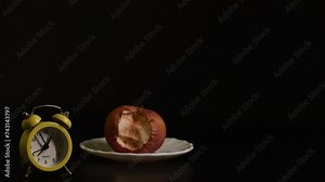 A yellow alarm clock shows the time against the background of a rotten apple on a saucer, black background. Passage of time, aging concept Stock Video