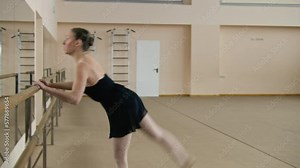 Female ballet dancer in training bodysuit stands near ballet barre, doing gymnastic leg movement, prepares for performance. Adult ballerina exercises in dance studio. School of classical ballet dance.