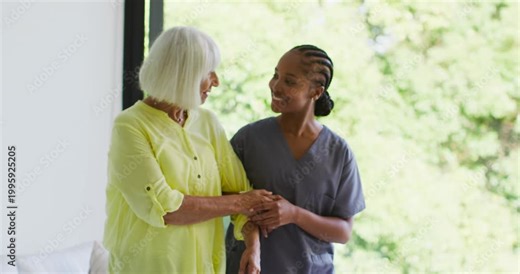 Female caregiver placing hand in scrubs guiding Senior woman taking steps for balance by glass door