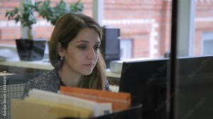 Female employee looks at computer screen while sitting at desk in office. Brunette reads attentively information on pc screen, propping face against hand. Young specialist, performs working tasks