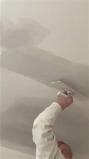 applying a smooth white plaster layer onto a high indoor ceiling surface using a square metal tool