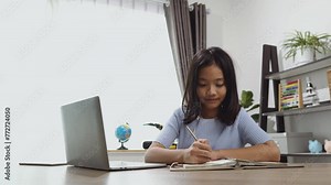 A little girl studies at a computer at home at a table with books and notebooks. Online training, distance learning, study online on computer.