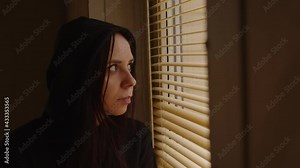 Close up of young woman watching through window blinds at home. Portrait of curious female observing through window jalousie in room.