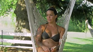 a young woman in a bikini swaying in a hammock while smoking a cigarette