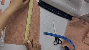 Seamstress's hands draw straight line on piece of fabric with ruler and pencil, then cut off excess canvas. Top view of designer's workshop with sewing machine and other tools. Craft and hobby.