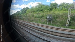 POV: Passenger view through the window while traveling by train across England. Public rail transport infrastructure with parallel railway lines under electric wires, surrounded by lush greenery.