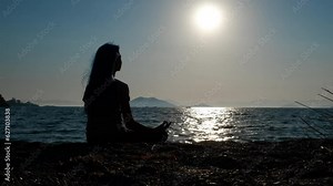 Silhouette teen meditates by sea. A view of girl silhouette sitting on lotus pose on the beach and admire the evening sun by the sea.