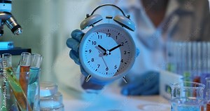 Scientist in laboratory holds an alarm clock and conducts experiments with colorful test tubes and samples on desk