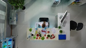 The frame above shows scientists sitting in a laboratory. A woman is doing research, testing, taking samples of vegetables that are near her. A man stands next to her and helps her. Top view
