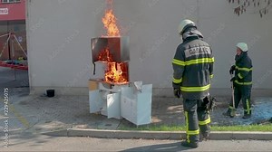 Firefighter pours more combustable liquid onto cabinets on fire to make it bigger for fire hose training as other fireman watches in foreground.