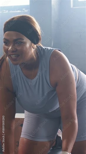 Vertical video: Bracing on plyo box, woman in blue set rowing hex dumbbell in gym building strength