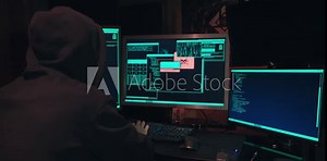 Hacker in a hood sits in front of computer screens and hacks databases. The concept of cyberterrorism, server hacking and cyberattacks. The room is dark, a person is typing code into the command line