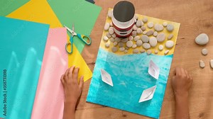 Kid hands make a lighthouse for school project at home. Top down view to wooden table with colored paper and other supplies