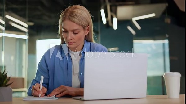 Concentrated blonde female using portable computer and notepad while sitting by personal table in coworking. Freelance video editor making schedule and using digital calendar and daybook indoor.
