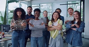 Front view of multi-ethnic group of people standing next to each other. Everyone crossing their hands and smiling while looking directly at camera. Posing for team photo. Office in background.