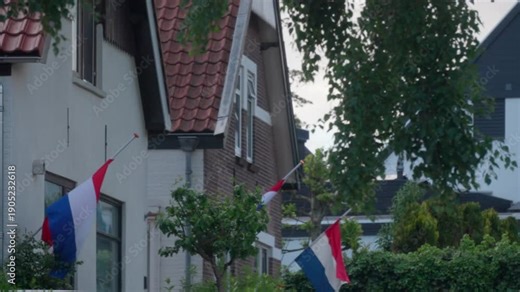 Perspective shot of multiple houses showing Dutch flags at half mast through green trees. Peaceful street scene during national day of mourning for war victims in the Netherlands.