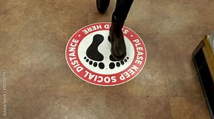 Due to ongoing COVID 19 pandemic US groceries, supermarkets and retail stores operate at limited capacity implement social distancing using floor signs. A customer steps on the designated spot.