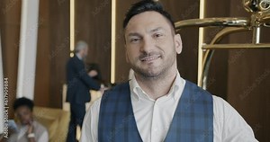 Smiling bellhop standing in hotel hall next to baggage cart and looking at camera. Guests are talking to receptionist in background.