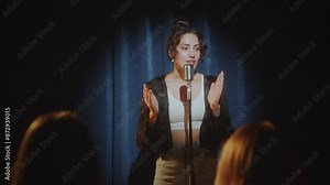 Young female stand-up comedian standing on stage in a spotlight and performing monologue in mic. View from audience Stock Video