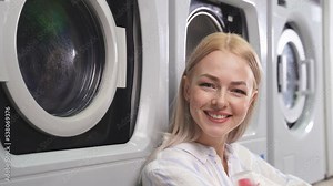 The washing machine of the public laundry washes dirty colored clothes, and an attractive woman sits by the car waiting for clean laundry