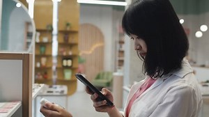 Side view of young Asian female customer scanning qr codes on skincare items while taking them from display and using mobile app for learning online information about products