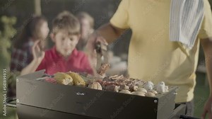 Man turning over meat on barbecue grill, his son blowing out fire. Father cooking roasted pork, mushroom and sweetcorn in garden, his family sitting in background. Barbecue with family concept
