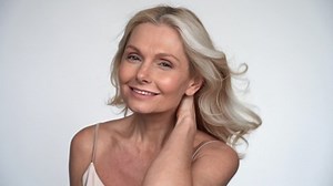 Happy smiling 50s middle aged mature woman with flying blond hair in blowing wind looking at camera. Old senior beauty model hairstyle, anti age shampoo for healthy hair care treatment, slow motion.