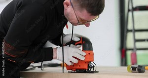 Man carpenter in safety glasses cuts wooden plank with jigsaw in workshop. Focused worker creates element for furniture using handy tool