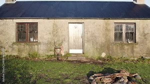 Aerial drone footage reversing directly away from the front door of a remote Scottish Bothy (Cadderlie Bothy in Glen Etive) over fields towards and facing a forested hillside in the morning light
